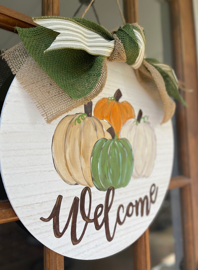 May include: A round wooden door hanger with a white background and a brown welcome sign. The door hanger is decorated with four painted pumpkins in shades of orange, white, green, and beige. The pumpkins are surrounded by a burlap bow with a green and white striped ribbon.