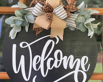 May include: A black round wooden door hanger with the word "Welcome" written in white cursive script. The hanger is decorated with a burlap bow and a leopard print ribbon, and greenery.
