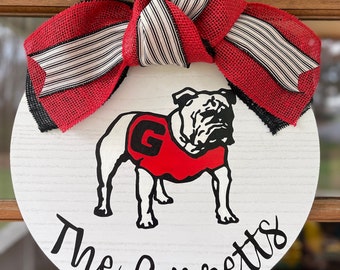 May include: A white round wooden door hanger with a red and black bow. The door hanger features a black and white illustration of a bulldog wearing a red jersey with a white "G" on it. The text "The Garretts" is written in black cursive below the bulldog.