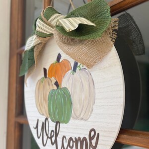 May include: A round wooden sign with a burlap and green ribbon bow. The sign is painted white with a wood grain texture and features a painted welcome message and four pumpkins in shades of orange, green, and white.