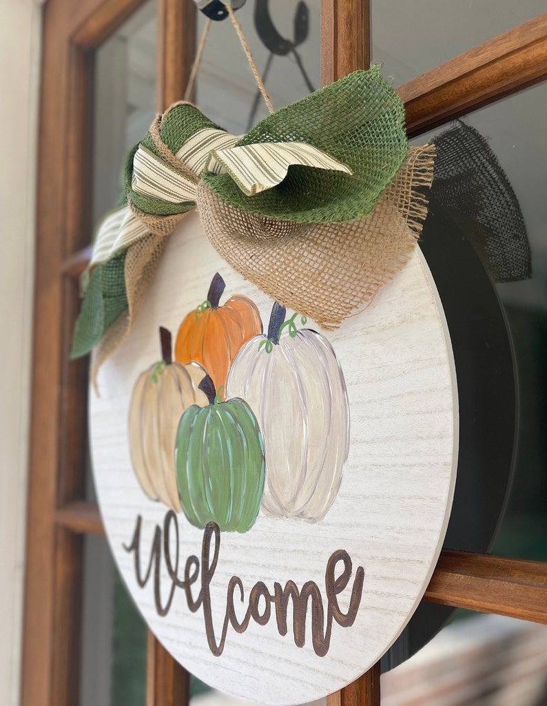 May include: A round wooden sign with a burlap and green ribbon bow. The sign is painted white with a wood grain texture and features a painted welcome message and four pumpkins in shades of orange, green, and white.