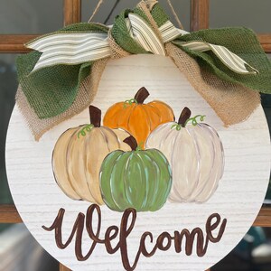 May include: A round wooden door hanger with a welcome sign painted in brown. The door hanger is decorated with a burlap bow and green and white striped ribbon. Four pumpkins are painted on the door hanger, one orange, one green, and two white.