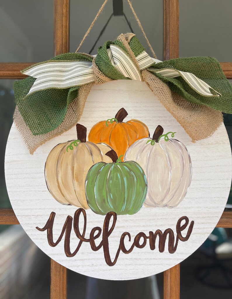 May include: A round wooden door hanger with a welcome sign painted in brown. The door hanger is decorated with a burlap bow and green and white striped ribbon. Four pumpkins are painted on the door hanger, one orange, one green, and two white.