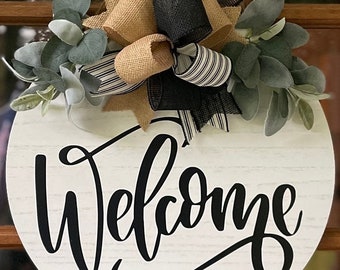 May include: A round white wooden sign with the word "Welcome" written in black cursive script. The sign is decorated with a burlap and black and white striped bow and greenery.