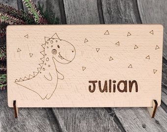 Może przedstawiać: Drewniany znak z kreskówkowym dinozaurem i wygrawerowanym imieniem Julian. Znak jest ozdobiony małymi trójkątami.
