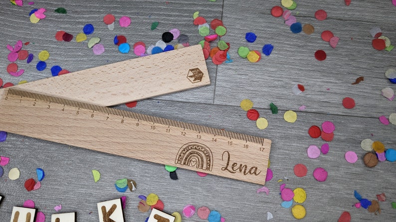 Może przedstawiać: Drewniana linijka z wygrawerowanym tęczą i imieniem "Lena". Linijka jest otoczona kolorowymi konfetti.