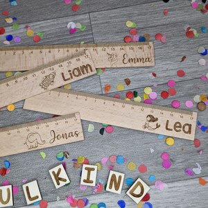 Może przedstawiać: Cztery drewniane linijki z r&oacute;żnymi ilustracjami i nazwami: Liam, Emma, Jonas i Lea. Linijki leżą na drewnianej powierzchni, wok&oacute;ł kt&oacute;rej rozrzucone są kolorowe konfetti. Słowo "KIND" jest ułożone z drewnianych liter.