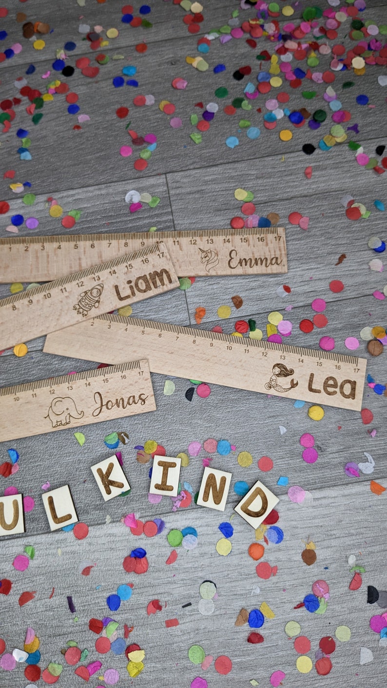 Może przedstawiać: Cztery drewniane linijki z r&oacute;żnymi ilustracjami i nazwami: Liam, Emma, Jonas i Lea. Linijki leżą na drewnianej powierzchni, wok&oacute;ł kt&oacute;rej rozrzucone są kolorowe konfetti. Słowo "KIND" jest ułożone z drewnianych liter.