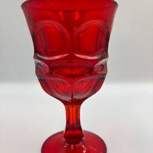 May include: A ruby red glass goblet with a decorative pattern. The goblet has a round base, a slender stem, and a bowl with a textured design. The glass is a vibrant, transparent red color.