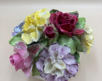 Könnte beinhalten: Ein dekoratives Blumenarrangement aus Porzellan mit einer Ansammlung skulptierter Blüten, darunter eine dunkelrote Rose, eine rosa Rose, gelbe Nelken und eine lavendelfarbene Nelke mit gerüschten Blütenblättern, akzentuiert durch kleine grüne Blätter.