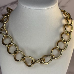 May include: A chunky gold-tone chain necklace displayed on a white mannequin bust. The necklace features large, interlocking oval links. The jewelry is a bold statement piece, perfect for adding a touch of glamour to any outfit.