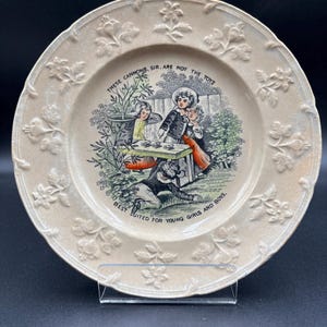 May include: Cream-colored ceramic plate with a detailed illustration of children playing, surrounded by floral relief patterns. The plate features text that reads "THESE CANNONS, SIR, ARE NOT THE TOYS BEST SUITED FOR YOUNG GIRLS AND BOYS."