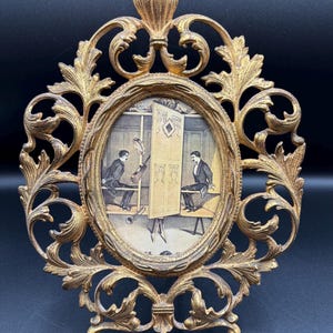 May include: Ornate gold-colored oval picture frame with a detailed filigree design. The frame surrounds a black and white print depicting two men in suits, one playing a musical instrument and the other seated. The frame has a decorative top and a base for standing.