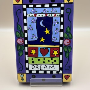 May include: A rectangular ceramic decorative piece with a purple background. The design includes a night sky scene with a crescent moon and stars, the word "DREAM", hearts, and floral accents. The piece is supported by a clear stand.