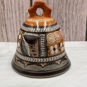 May include: Hand-painted ceramic bell with a brown, orange, and tan color scheme. The bell features decorative floral and geometric patterns in white and black. The bell has a loop at the top for hanging.
