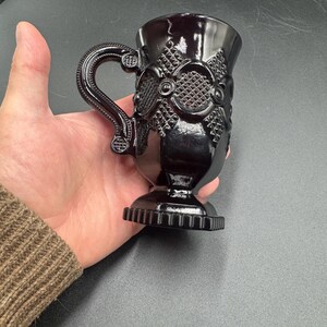 May include: A dark purple glass mug with an ornate handle and detailed embossed patterns. The mug has a pedestal base with a ridged design. The mug is held in a hand against a dark background.