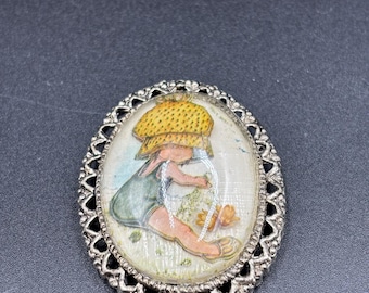 May include: Vintage oval brooch with a silver-tone filigree frame. The brooch features a painted image of a child wearing a yellow bonnet and green overalls. The child is sitting in a field of flowers. A charming piece of vintage jewelry.