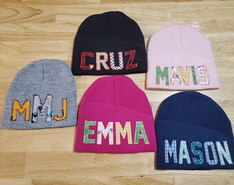 May include: Five knitted hats with different names embroidered on them. The names are "MMJ", "CRUZ", "MAVIS", "EMMA", and "MASON".