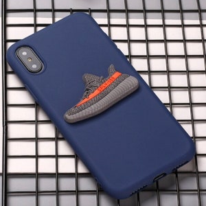 Puede incluir: Una funda de tel&eacute;fono azul marino con una zapatilla deportiva 3D gris y naranja unida a la parte posterior. La zapatilla tiene el texto "SPLY-350" en el lateral.