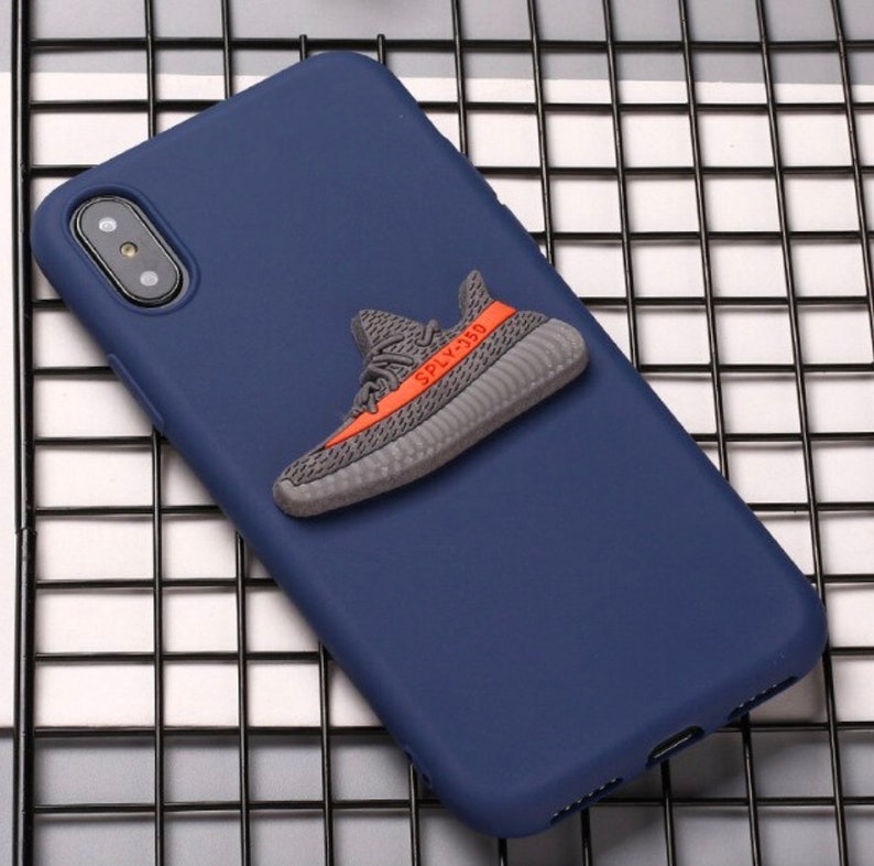 Puede incluir: Una funda de tel&eacute;fono azul marino con una zapatilla deportiva 3D gris y naranja unida a la parte posterior. La zapatilla tiene el texto "SPLY-350" en el lateral.