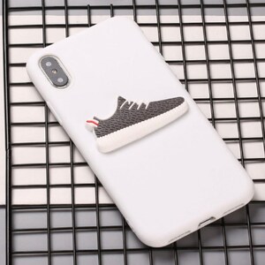 Puede incluir: Funda de tel&eacute;fono blanca con una zapatilla 3D gris y blanca unida a la parte posterior.