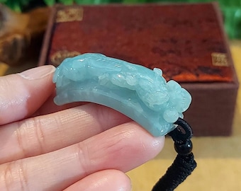 Puede incluir: Un colgante de jade verde claro tallado con un diseño de dragón, colgado de un cordón negro con una cuenta negra.