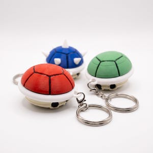 May include: Three turtle shell keychains featuring a red shell, a green shell, and a blue spiked shell. Each keychain has a white base, black detailing, and a silver metal ring attached to a small loop on the side of the shell structure.