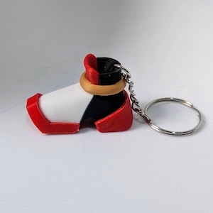 Puede incluir: Un llavero de bota impreso en 3D de color rojo, blanco y negro con un anillo dorado y una cadena plateada.