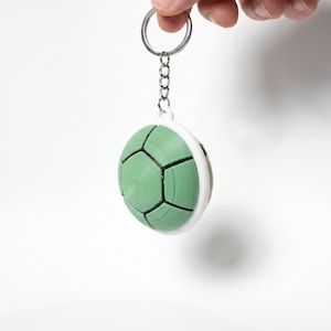 May include: A keychain featuring a spherical, light green turtle shell charm with black geometric lines. The charm is attached to a silver-toned metal split ring by a short chain. The shell has a smooth, rounded surface and a white border.