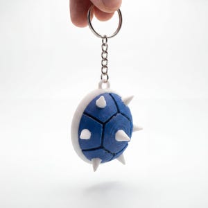 May include: A blue and white spiky shell keyring featuring a circular blue centre with white hexagonal borders and protruding white spikes. The shell is attached to a silver-coloured metal split ring by a short metal chain link connector.