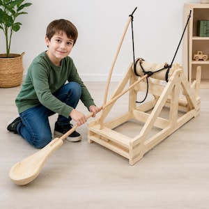 Puede incluir: Un juguete de catapulta hecho de madera natural de color claro, con un marco, un eje central giratorio y un brazo lanzador largo con un extremo en forma de cuchara. El mecanismo está conectado por una cuerda negra para su uso manual.