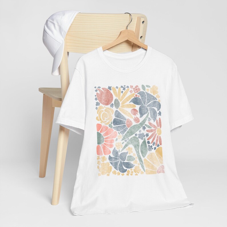 May include: White t-shirt with a colorful floral print design. The print features a variety of flowers and leaves in shades of pink, blue, yellow, and green.