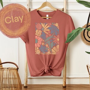 May include: A clay-colored t-shirt with a floral print in shades of blue, orange, yellow, and green. The print is a square design with a variety of flowers and leaves.