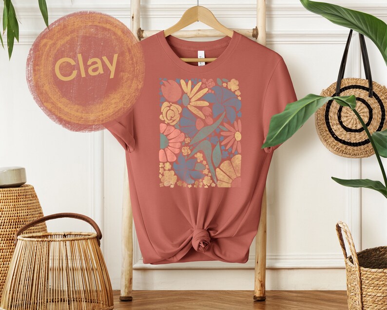 May include: A clay-colored t-shirt with a floral print in shades of blue, orange, yellow, and green. The print is a square design with a variety of flowers and leaves.