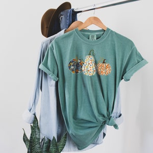 May include: A teal green t-shirt with a graphic of three pumpkins. The pumpkins are patterned with floral designs in shades of orange, blue, and yellow.