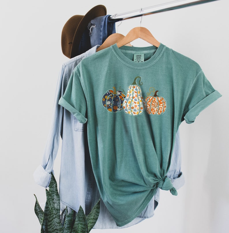 May include: A teal green t-shirt with a graphic of three pumpkins. The pumpkins are patterned with floral designs in shades of orange, blue, and yellow.