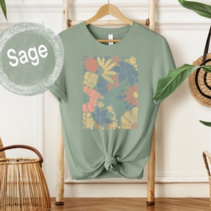 May include: Sage green t-shirt with a floral print featuring a variety of colors, including pink, yellow, blue, and orange.