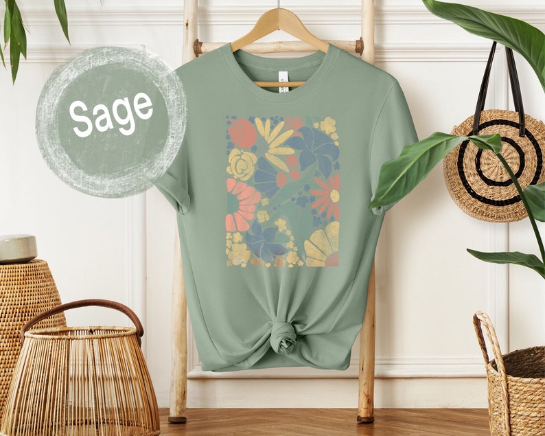 May include: Sage green t-shirt with a floral print featuring a variety of colors, including pink, yellow, blue, and orange.