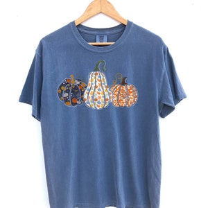 May include: A blue t-shirt with a graphic of three pumpkins. The pumpkins are patterned with different floral designs in shades of orange, yellow, and blue.