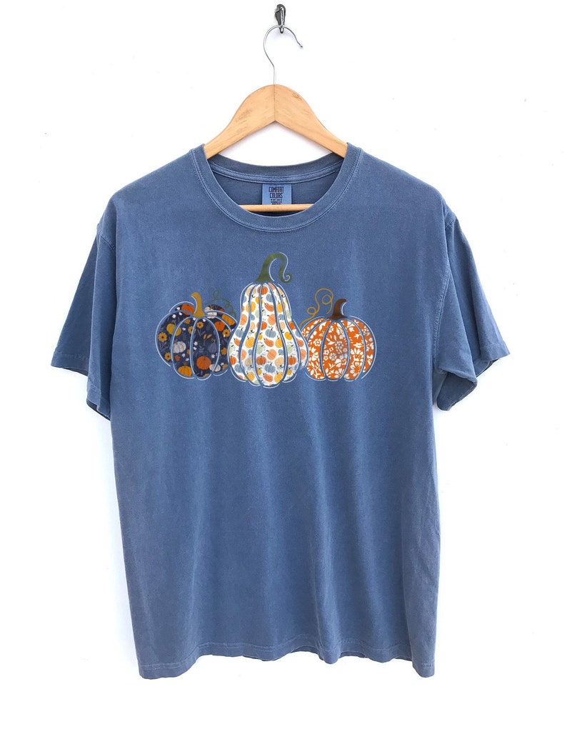 May include: A blue t-shirt with a graphic of three pumpkins. The pumpkins are patterned with different floral designs in shades of orange, yellow, and blue.