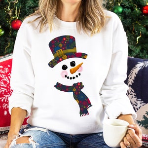 Puede incluir: Sudadera blanca con un dise&ntilde;o de mu&ntilde;eco de nieve floral colorido. El mu&ntilde;eco de nieve tiene un sombrero negro, una nariz de zanahoria y una bufanda con estampado floral.