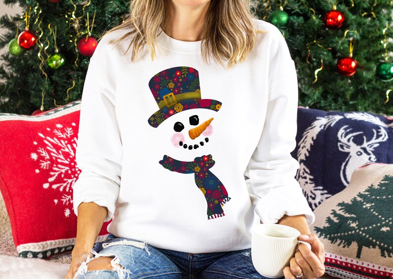 Puede incluir: Sudadera blanca con un dise&ntilde;o de mu&ntilde;eco de nieve floral colorido. El mu&ntilde;eco de nieve tiene un sombrero negro, una nariz de zanahoria y una bufanda con estampado floral.