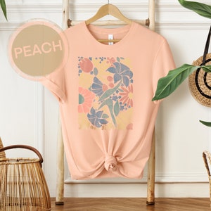 May include: Peach colored t-shirt with a floral print design. The print features a variety of flowers in shades of pink, blue, and yellow.