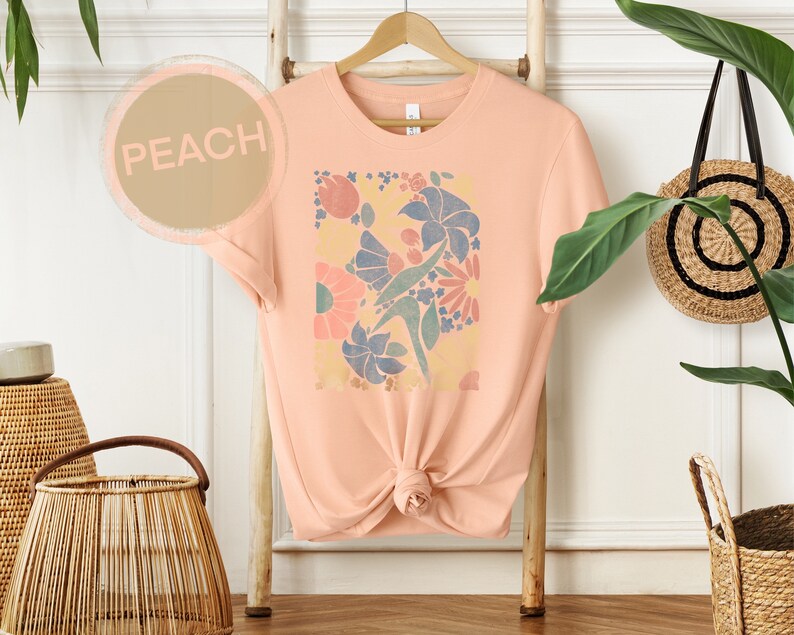 May include: Peach colored t-shirt with a floral print design. The print features a variety of flowers in shades of pink, blue, and yellow.
