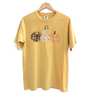 May include: A yellow short-sleeved t-shirt with a graphic of three pumpkins. The pumpkins are patterned with floral designs in shades of orange, blue, and green.