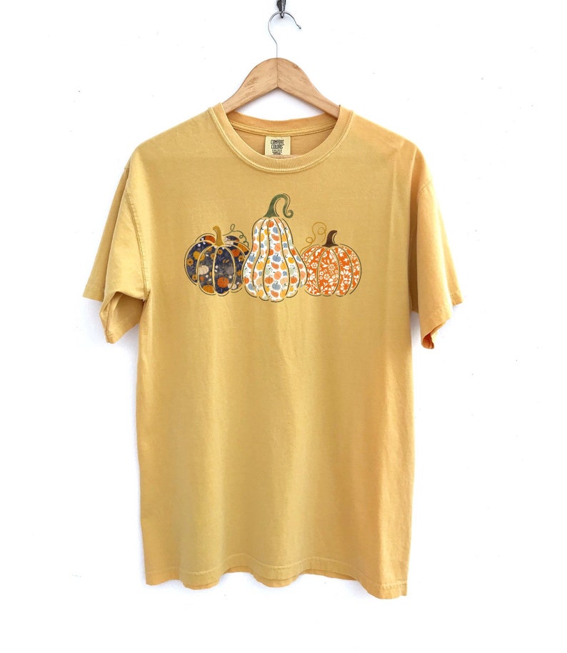 May include: A yellow short-sleeved t-shirt with a graphic of three pumpkins. The pumpkins are patterned with floral designs in shades of orange, blue, and green.
