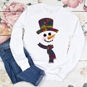 Puede incluir: Camiseta blanca de manga larga con un dise&ntilde;o de mu&ntilde;eco de nieve con estampado floral. El mu&ntilde;eco de nieve tiene un sombrero de copa negro, ojos negros, una nariz de zanahoria y una bufanda floral roja y azul.