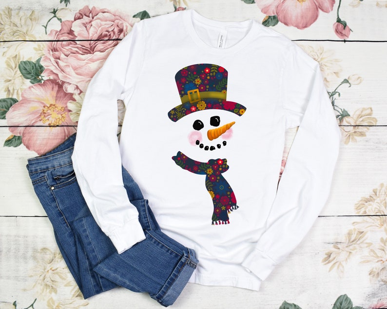 Puede incluir: Camiseta blanca de manga larga con un dise&ntilde;o de mu&ntilde;eco de nieve con estampado floral. El mu&ntilde;eco de nieve tiene un sombrero de copa negro, ojos negros, una nariz de zanahoria y una bufanda floral roja y azul.