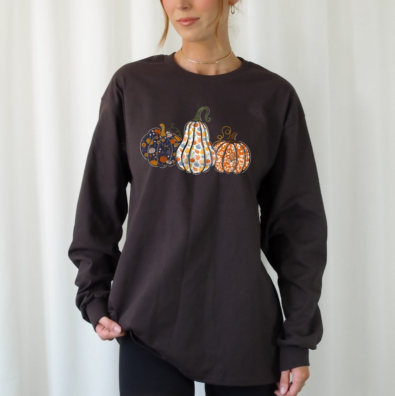 May include: A dark brown long-sleeved shirt with a graphic of three pumpkins. The pumpkins are decorated with floral patterns in shades of orange, white, and blue.
