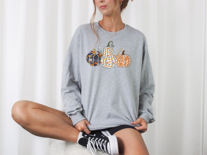 May include: A gray long-sleeved sweatshirt with a design of three pumpkins. The pumpkins are patterned with different colors and designs.